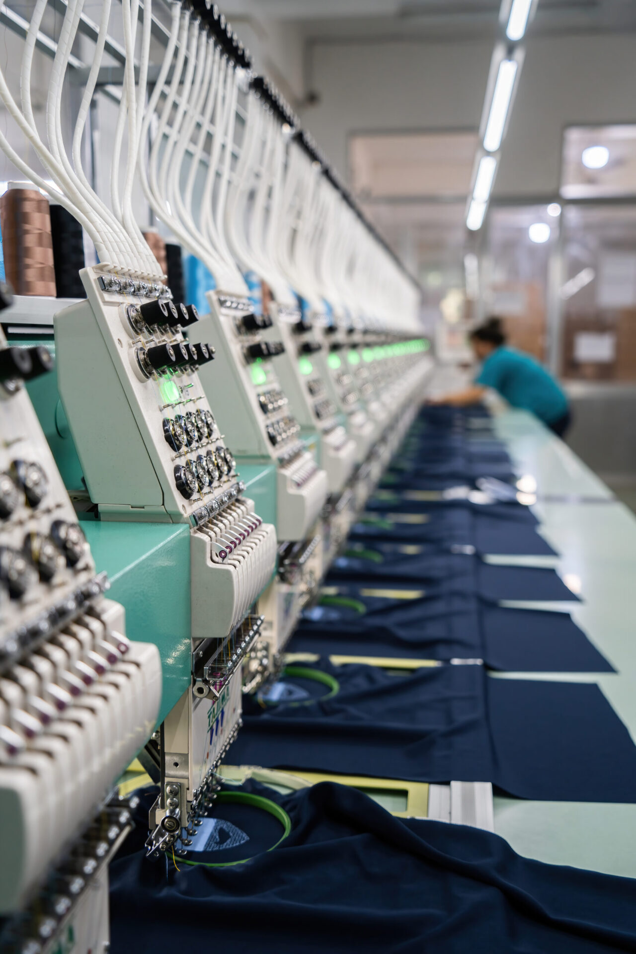 Long row of industrial commercial embroidery machines with multiple thread spools running simultaneously to stitch custom logos onto navy garments in a professional production facility, demonstrating the large-scale capacity for custom embroidery services available for bulk branded apparel orders at The Logo Store in Austin TX