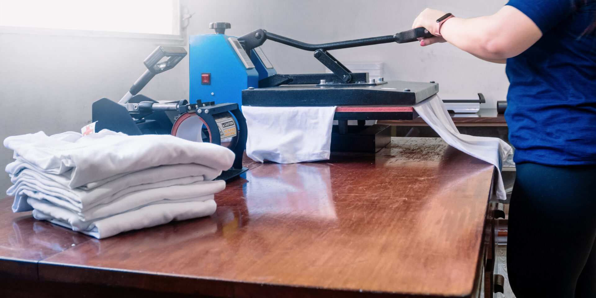 Print technician operating a blue heat press machine while positioning a white t-shirt on the printing pad with a stack of blank shirts ready for production, demonstrating the professional custom heat press printing services for apparel orders at The Logo Store in Austin TX Image Title: Custom Heat Press Printing Services Austin TX | T-Shirt Production | The Logo Store