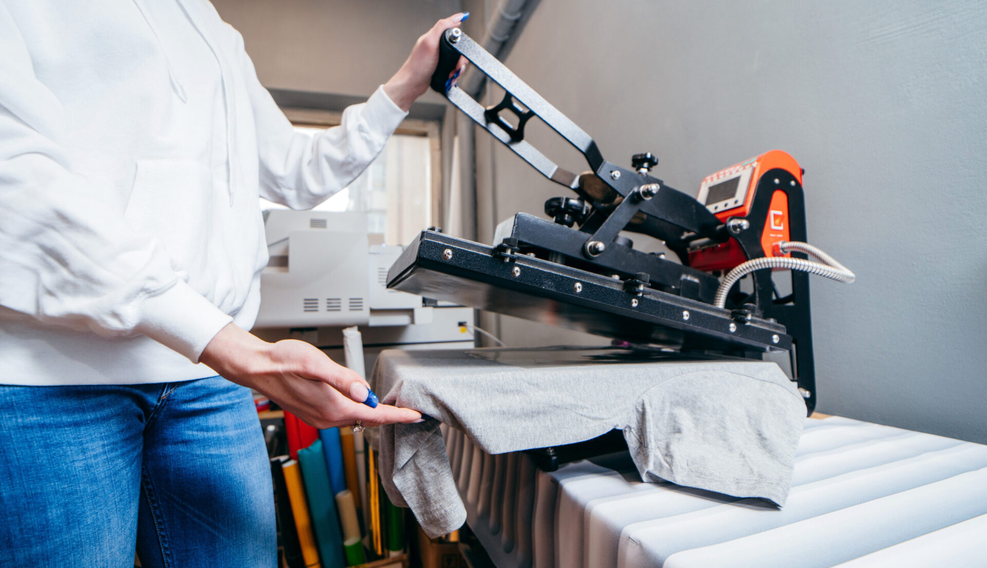 Small business owner lifting the platen of a heat press machine after transferring a custom design onto a gray t-shirt, demonstrating heat press printing as a low-cost, low-minimum option for how to start a clothing brand in 2026
