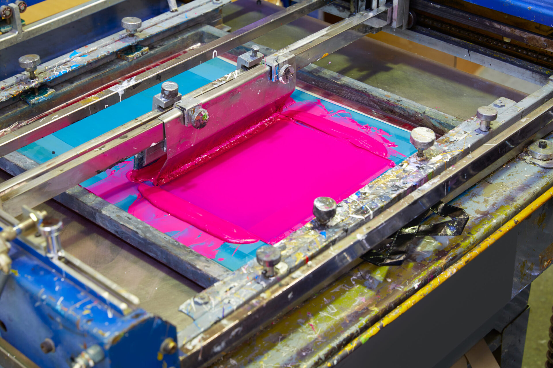 Close-up of vibrant pink ink loaded onto a blue mesh screen printing frame on a professional press, showing the bold color output available with no minimum custom shirt printing at The Logo Store in Austin TX