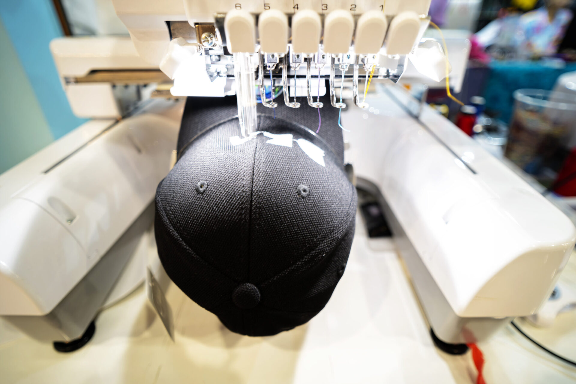 Commercial embroidery machine with multiple numbered needle heads stitching a custom logo onto a structured black baseball cap, demonstrating why embroidery is the preferred method over screen printing for custom branded hats and headwear at The Logo Store in Austin TX