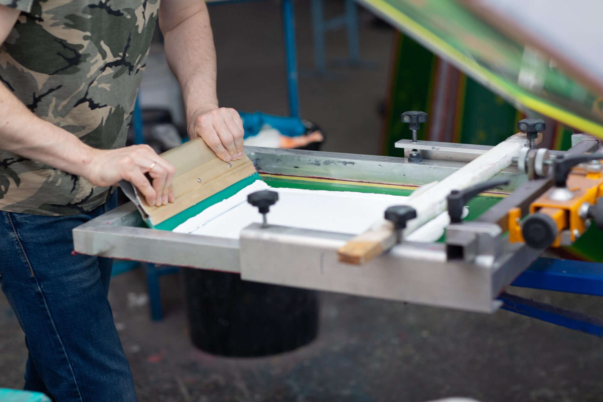 Print technician using a wooden squeegee to pull ink across a green mesh screen on a professional screen printing press, illustrating how screen printing works compared to embroidery for custom apparel orders at The Logo Store in Austin TX