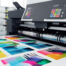 A large format digital printing machine producing vibrant full-color prints showing the range of color options available when choosing the best printing method for custom t-shirts and branded apparel at The Logo Store in Austin TX