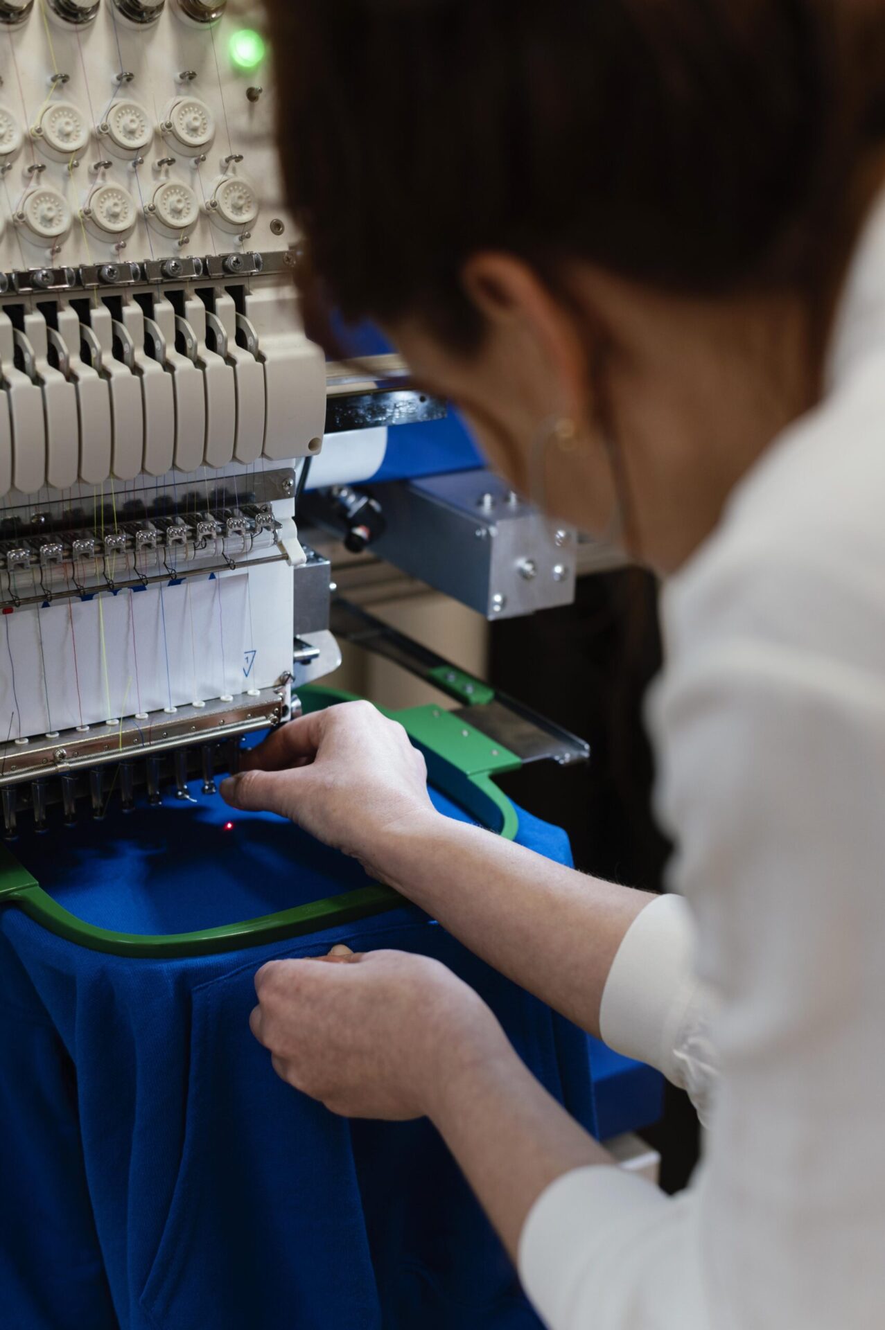Technician adjusting fabric on a commercial embroidery machine with multiple thread spools stitching a custom design onto a blue garment, showcasing embroidery as a premium printing method option for custom t-shirts and branded apparel at The Logo Store in Austin TX