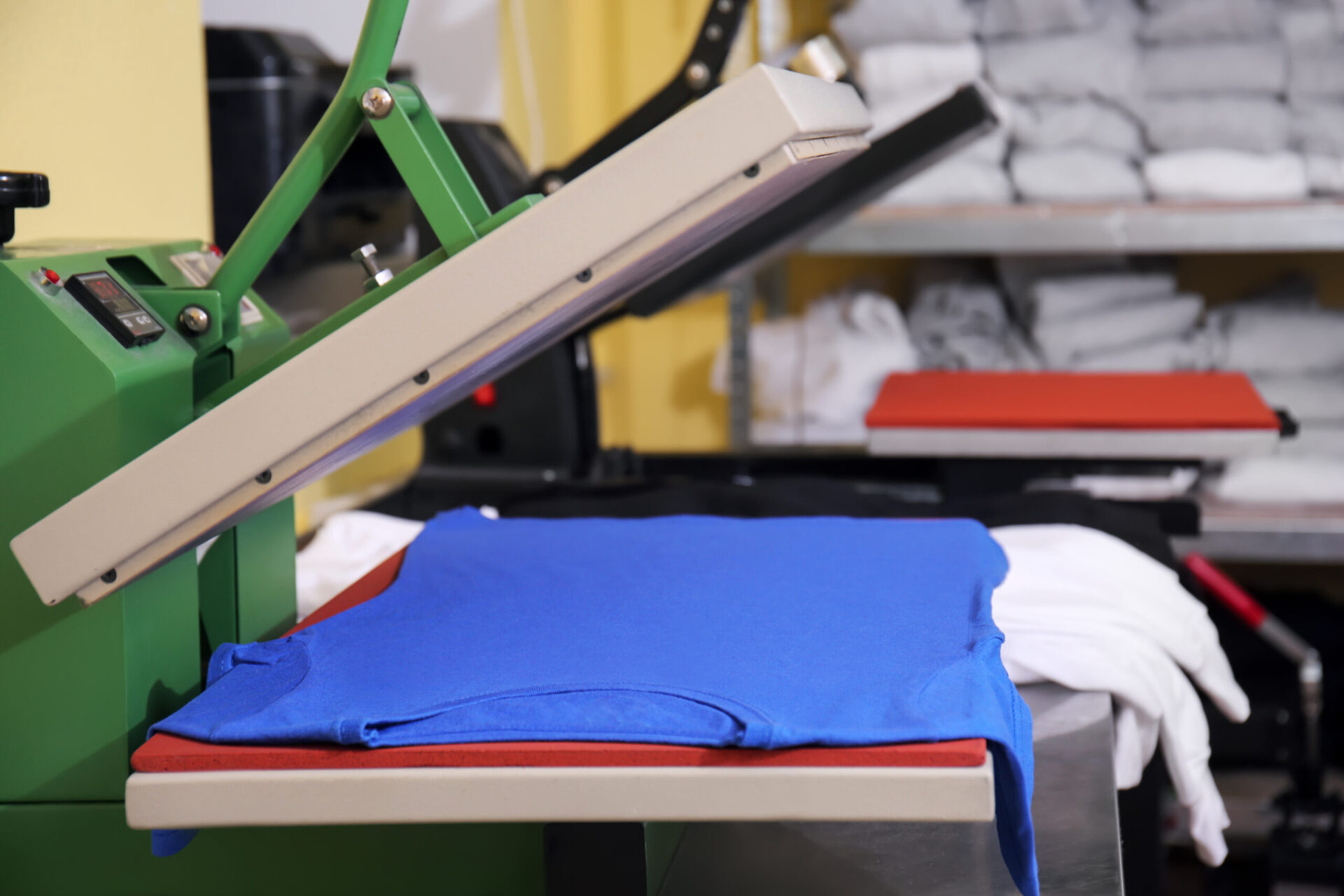 A heat press machine with its platen raised revealing a blue t-shirt positioned on the printing pad, demonstrating the heat press printing method as one of the best options for custom t-shirts with complex designs or small batch orders at The Logo Store in Austin TX