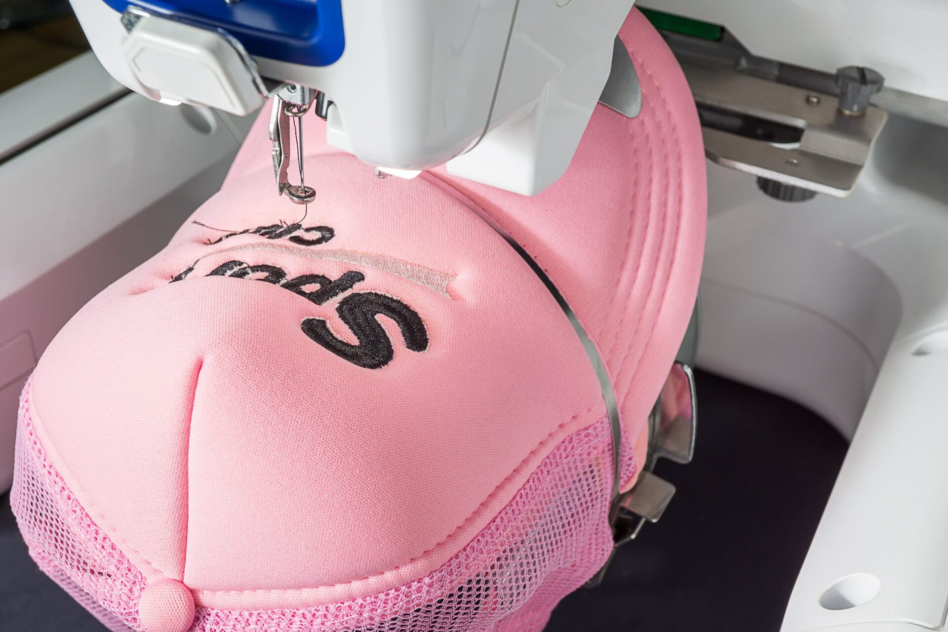 Commercial embroidery machine stitching bold black custom text onto a structured pink trucker hat, showing how custom hat embroidery works for branded caps and headwear orders at The Logo Store in Austin TX