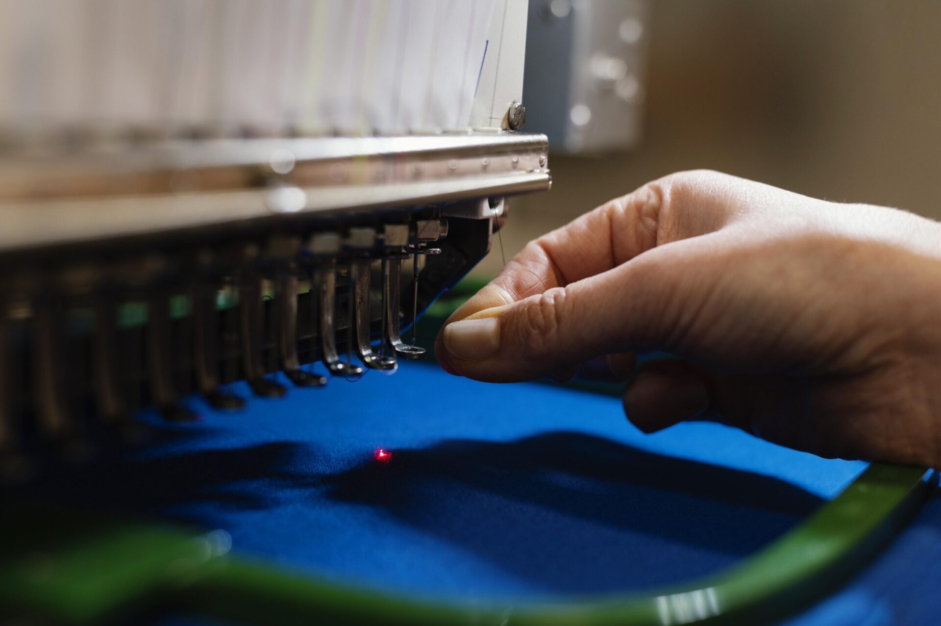 Technician guiding blue fabric under a commercial embroidery machine with a red laser guide point, demonstrating the precision process of custom embroidery for branded hoodies and apparel orders at The Logo Store in Austin TX