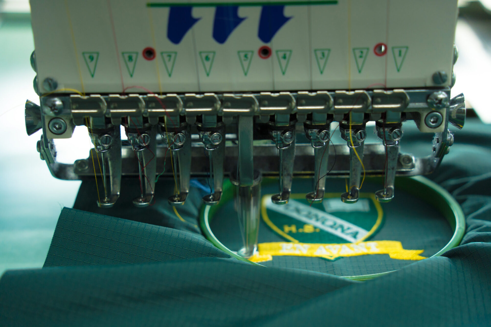 Commercial multi-needle embroidery machine stitching a detailed green and yellow logo design onto a teal polo shirt, showing how custom embroidery works for branded team uniforms and corporate apparel at The Logo Store in Austin TX