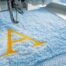 Close-up of a commercial embroidery machine needle stitching a bold gold letter A design onto soft blue fabric, visually explaining what embroidery is and how the thread-based decoration process works for custom branded apparel at The Logo Store in Austin TX
