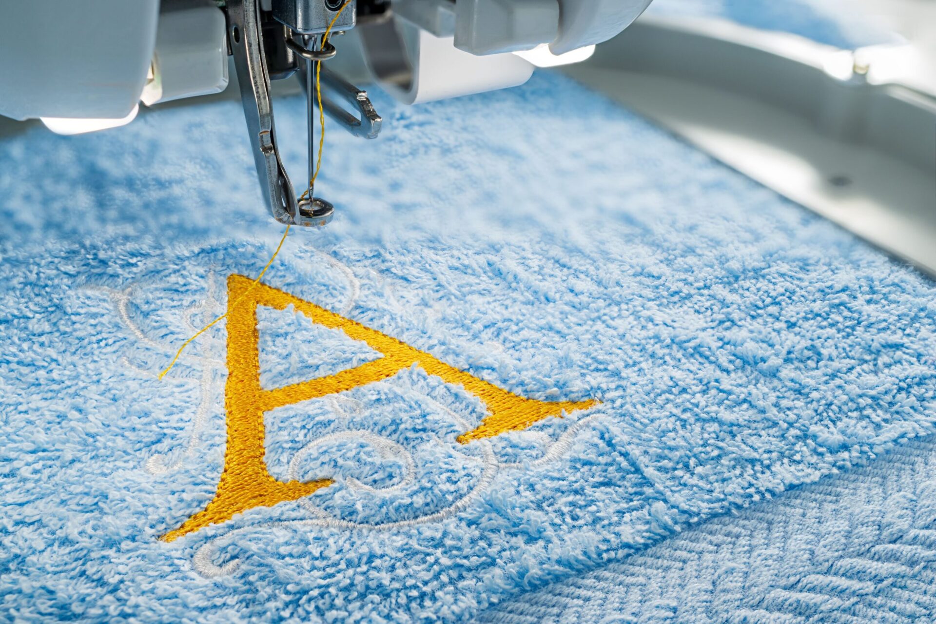 Close-up of a commercial embroidery machine needle stitching a bold gold letter A design onto soft blue fabric, visually explaining what embroidery is and how the thread-based decoration process works for custom branded apparel at The Logo Store in Austin TX