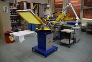 Professional multi-station screen printing machine with yellow mesh screens set up in a custom print shop, showing the automated equipment used for bulk t-shirt and apparel screen printing orders