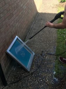 Screen printing technician washing out hardened emulsion from a mesh screen with a pressure washer, revealing the stencil design after UV exposure as part of the screen preparation process for custom apparel printing