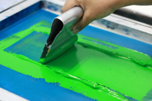 Close-up of a squeegee pushing bright green ink across a silk screen during the screen printing process, showing how ink is evenly distributed through the mesh stencil to create custom printed apparel